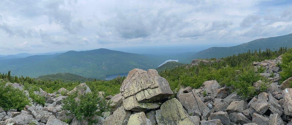 Mountaintop view with large boulders in the foreground and green mountain peaks in the background. In the middle, below, you can see a small lake peeking out. The sky is hazy with clouds and sun.