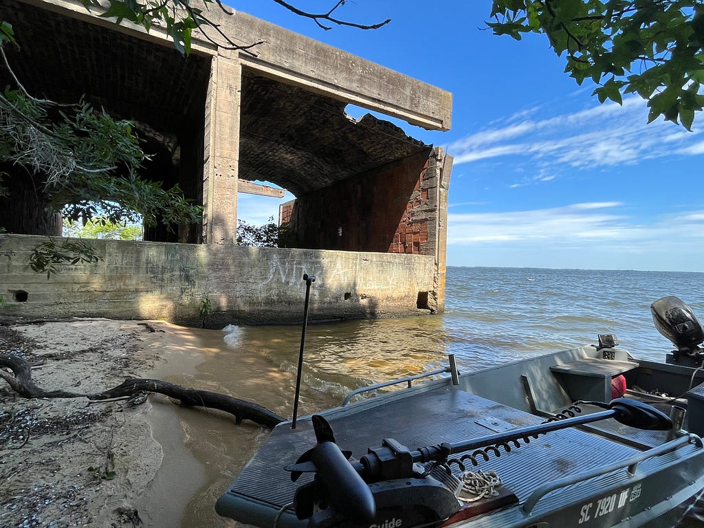 An image of a building, formerly a kiln, abandoned. There is a boat in front of the kiln, the building is now mostly bricks and concrete foundation. Water surrounds the kiln, and submerges the building. A tree juts out in the image from the left hand side.