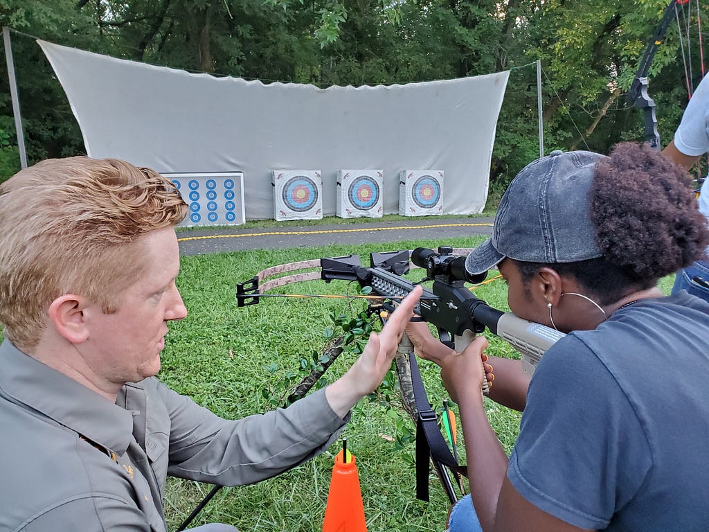 A young woman holds up a crossbow aimed at archery targets with the guidance of a man training her
