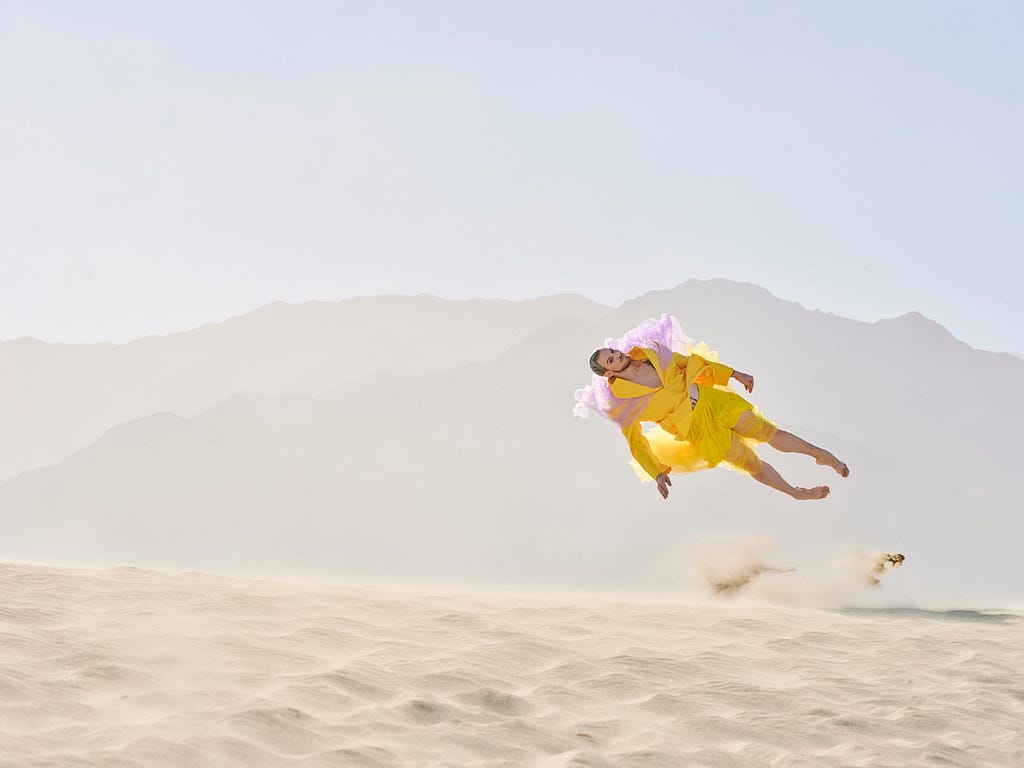 Young man in fashionable, yellow outfit falling onto sand in desert