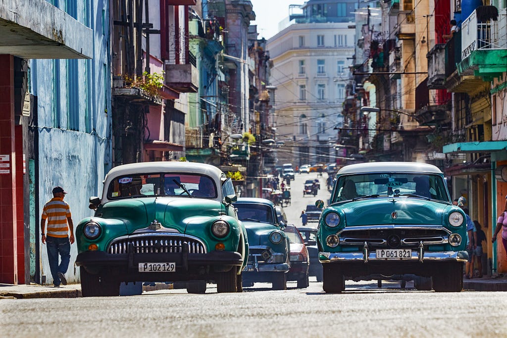 2 Vintage  cars. 1 turquoise with white roof, other light green with white roof in sunlit centre of busy Havana, Cuba.