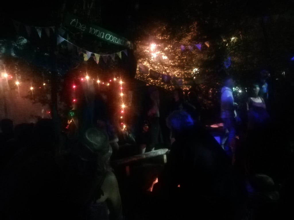 A woodland scene at night with fairy lights and bunting and some people. A sign saying ‘The Iona Community’ hangs at the top.