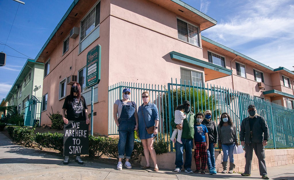 9 tenants stand in front of their building. One tenant holds a sign that says “WE ARE HERE TO STAY”