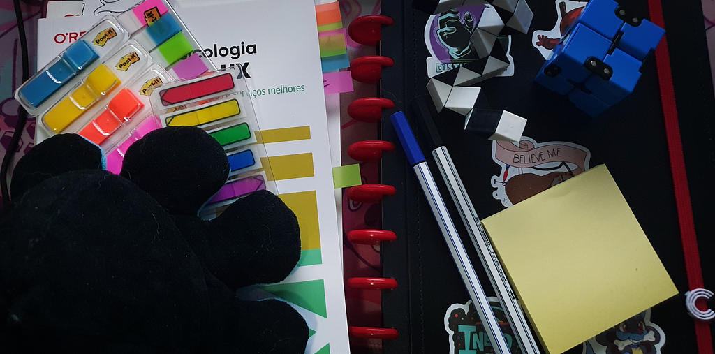 Foto de itens que a autora usa no seu dia a dia: um caderno com capa preta, uma caneta azul, uma caneta preta, um bloco adesivo amarelo, um polvo de pelúcia preto, fitas adesivas coloridas e um cubo mágico azul