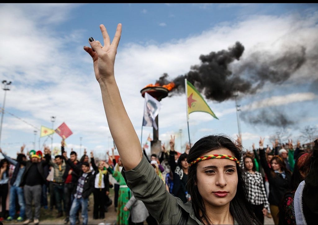 A Kurdish woman raises her hand in the air to make the peace sign. She is wearing a braided headband in the national colours. In the background are a crowd of Kurdish people waving flags and letting off smoke signals.