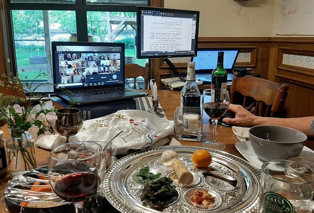 A traditional seder plate with two computer screens above it, one with a Zoom call meeting, another with a Haggadah page.