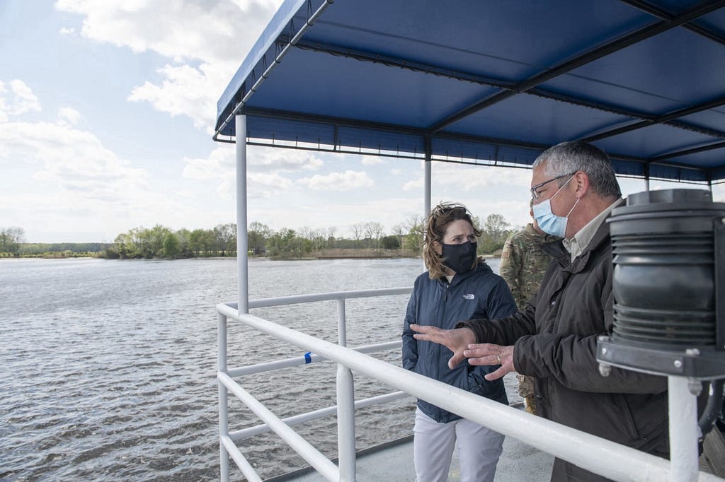 A woman and man stand talking on a boat that looks like a small ferry. Both are wearing protective masks. The river is visible behind them, and behind that the marsh horizon line.