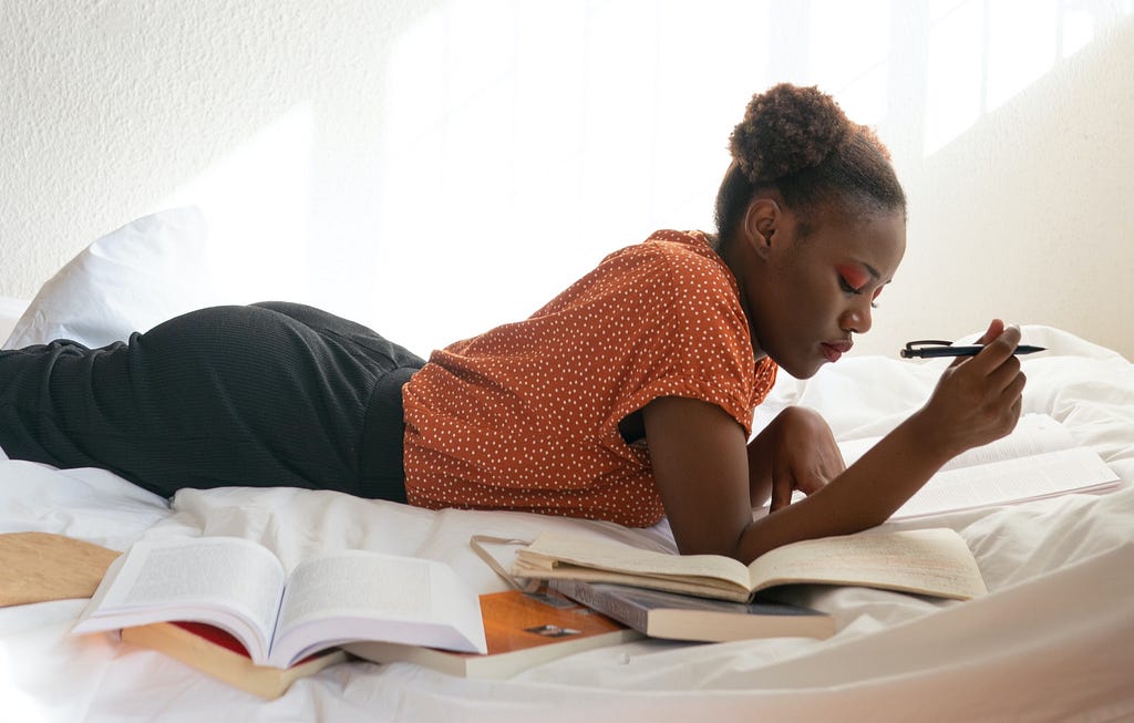 Black female presenting person laying across a bed reading several books and taking notes.