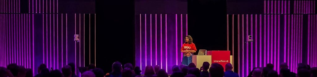 For everyone to see: A woman having a keynote on a stage of a conference with a big audience in the foreground of the picture. It is a dark space with stage lighting and a purple lighted background.