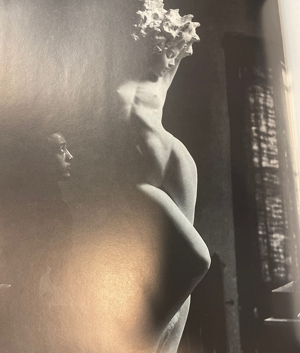 Isamu Noguchi looks at a sculpture of a naked woman — possibly a goddess — in a sensual pose. Black and white image.