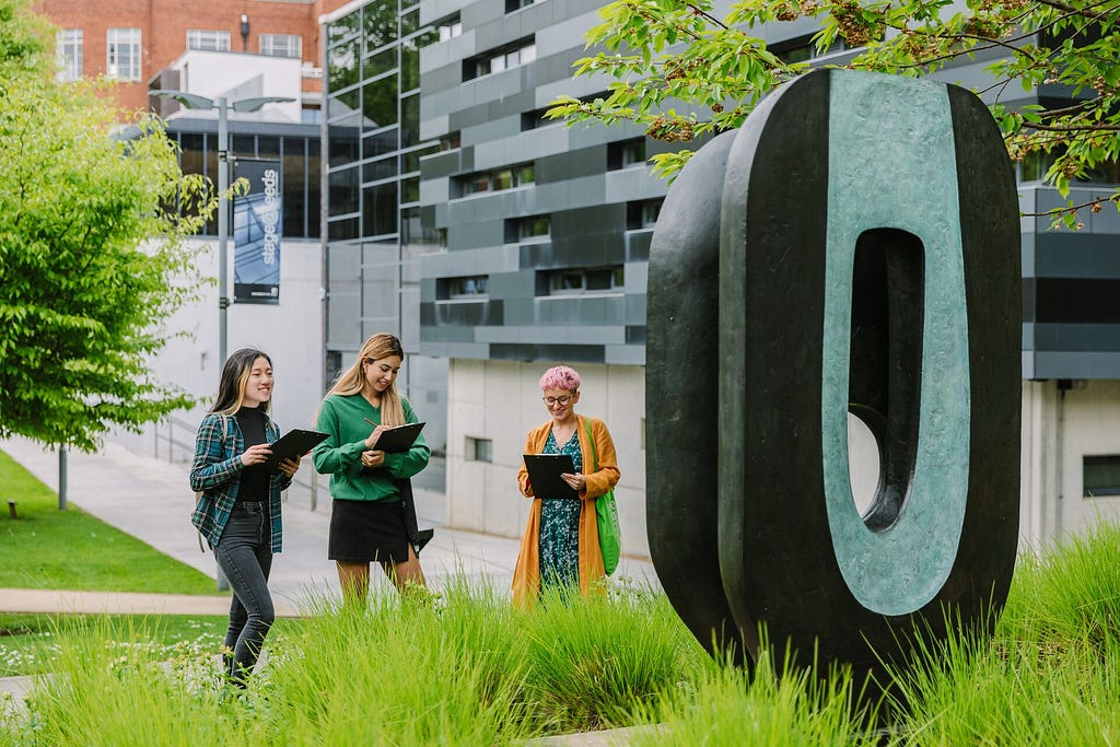Three students sketch Barbara Hepworth’s ‘Dual Form’ sculpture, an abstract bronze piece, located on the Leeds University campus as part of the Public Art Trail. The sculpture stands among greenery, with modern campus buildings in the background.