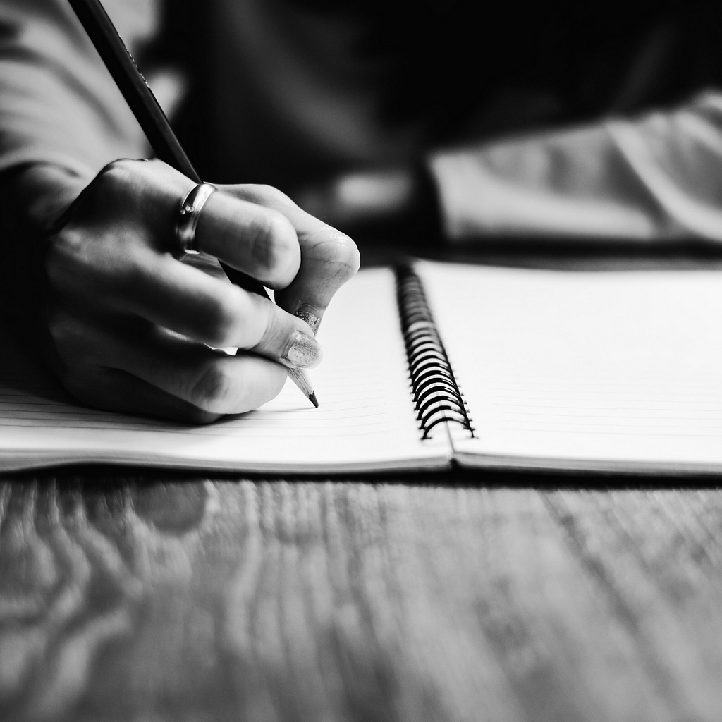 a person’s hand holding a pencil and writing in a spiral-bound notebook