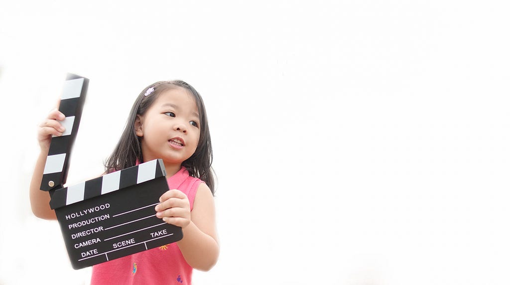 A young Asian girl holds a black and white film clapper and looks off to the right of the frame.
