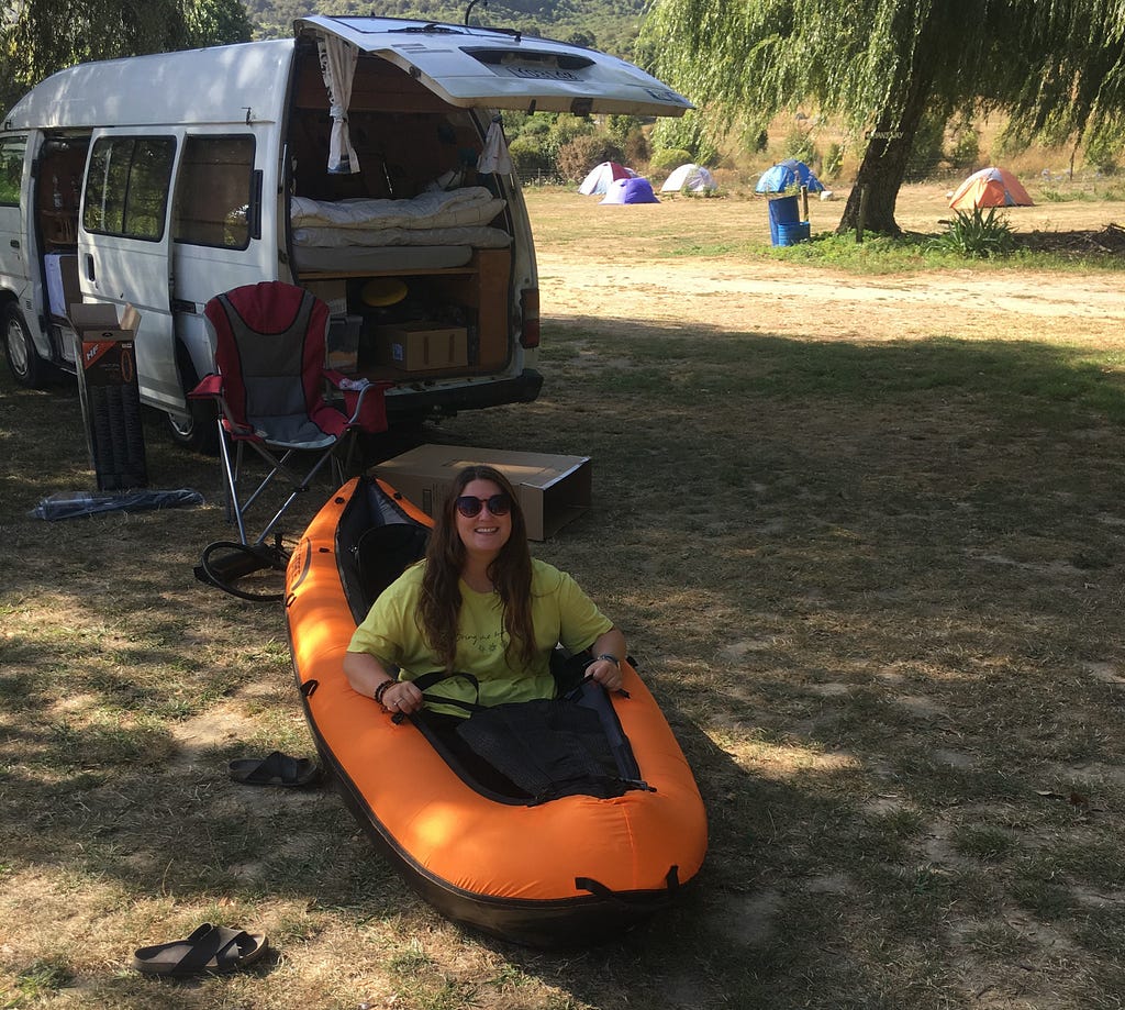 A girl with dark hair, wearing a yellow t-shirt and sunglasses sitting in an orange inflatable kayak on patchy grass.