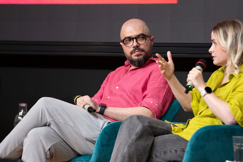 Two people seated in chairs and in the middle of a discussion, with one person speaking into a microphone and the other person listening.