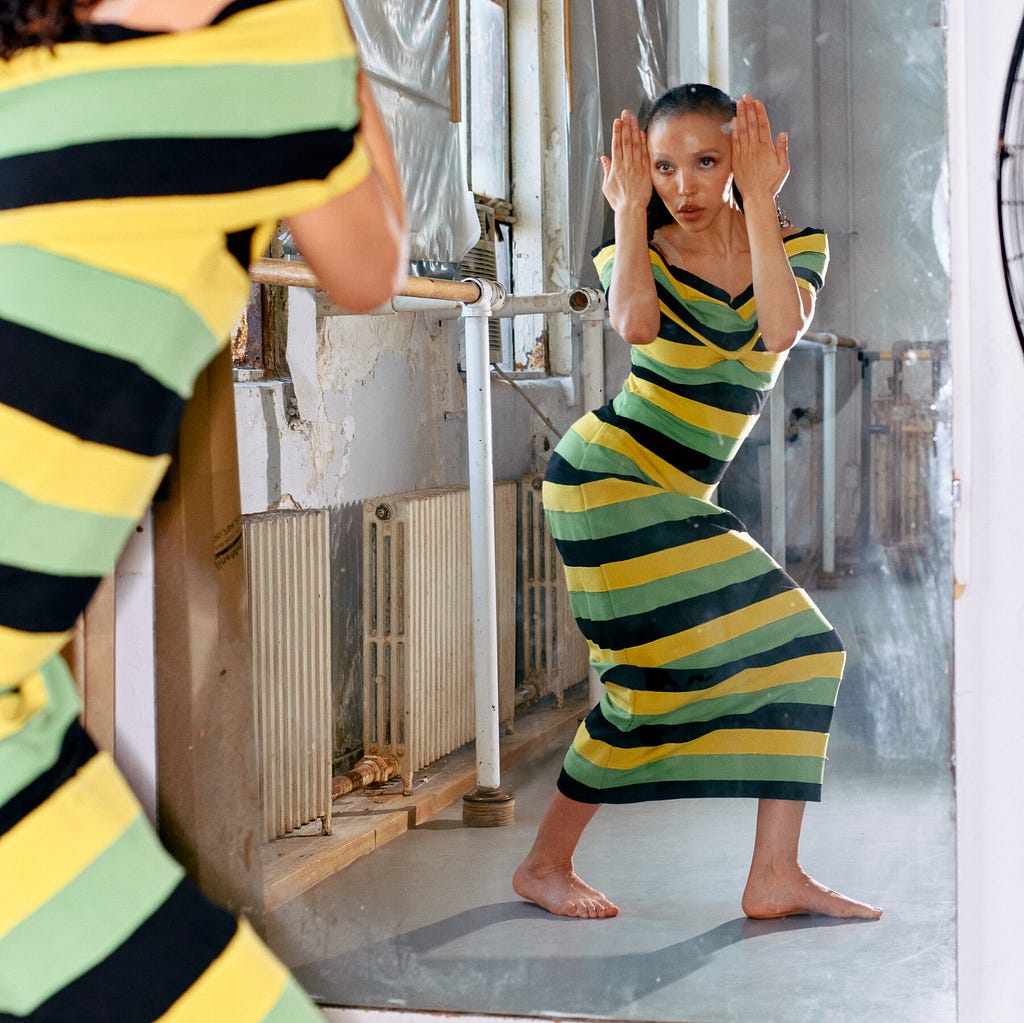 Artist F.K.A Twigs is seen posing in front of a mirror in the Martha Graham Studios wearing a black, green, and yellow striped body fitting dress.