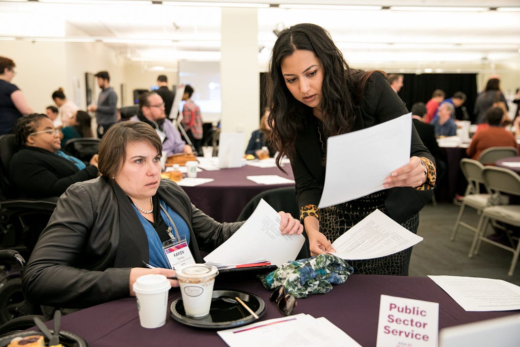 Azeema Akram holds up a paper document for Karen Tamley to examine. Karen sits at a table, indoors, at an ADA 25 event.