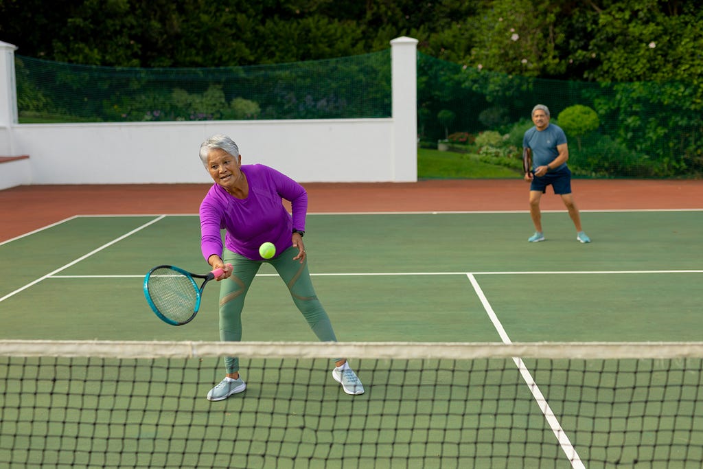 Photo of a male and female couple playing doubles tennis. She is at the net taking a low volley. The couple have olive skin and are in their mid to late 60s.