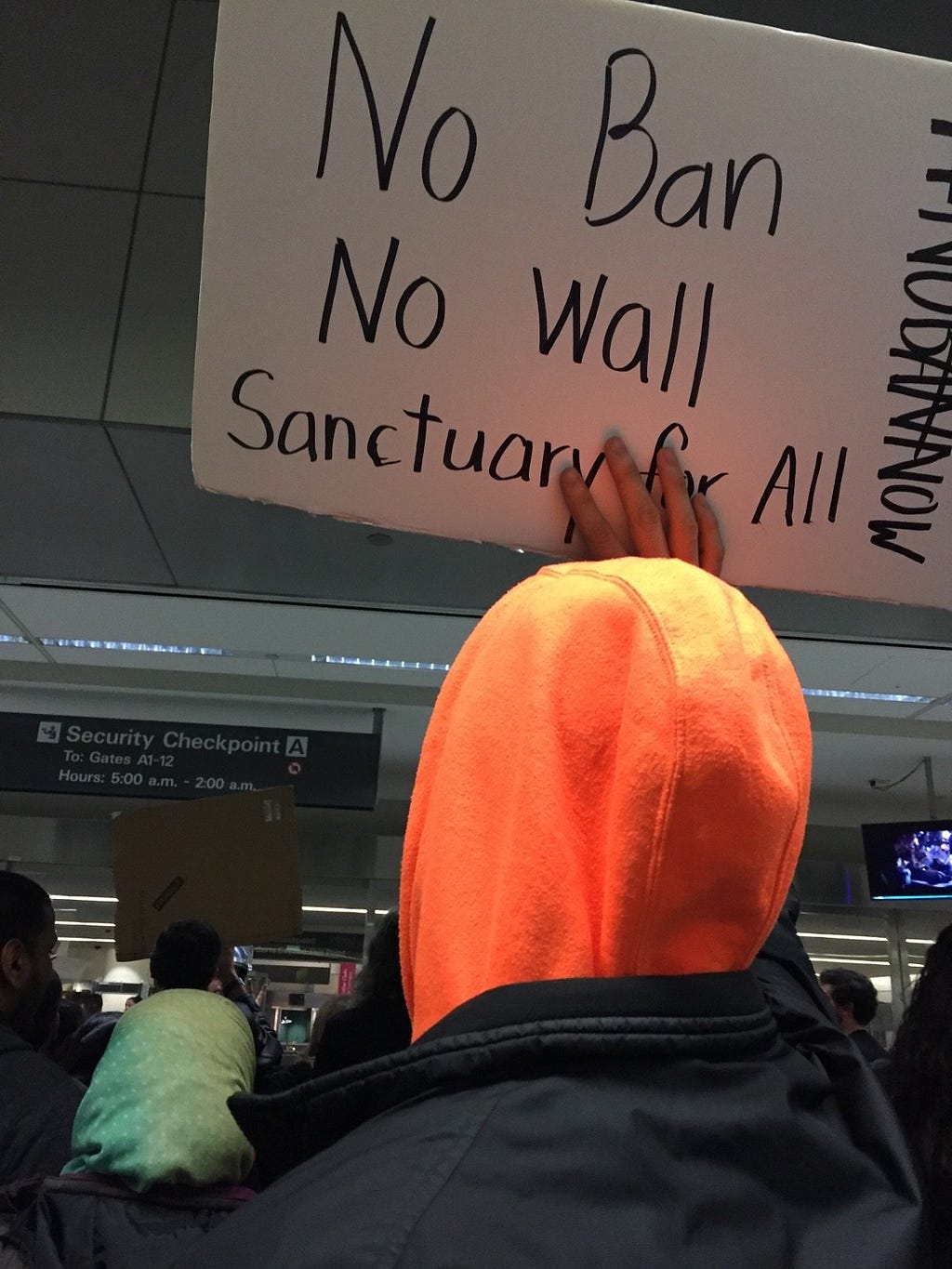 A brown person holds up a sign in a busy airport lobby that says “No Ban No Wall Sanctuary for All.”