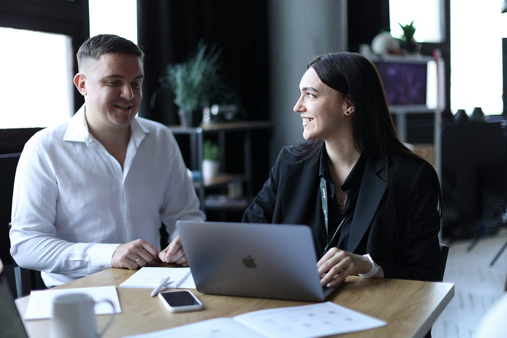 Two people in an office sitting and smiling at one another in front of a computer.