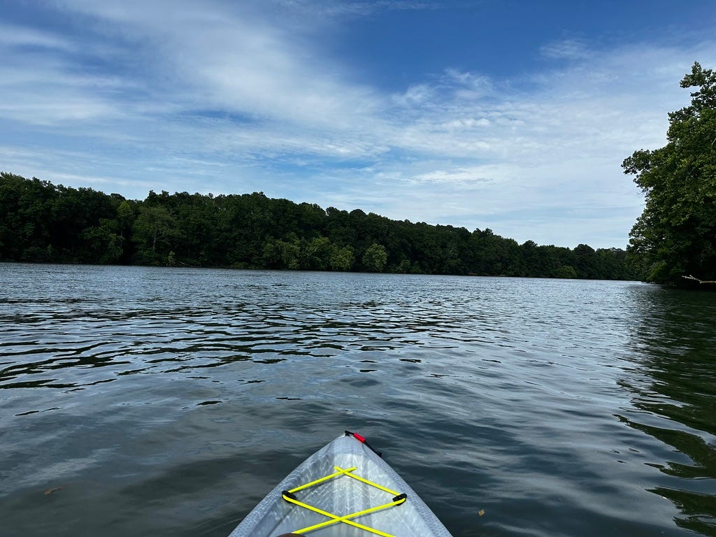 Front of grey kayak on wide river with tress lining sides.