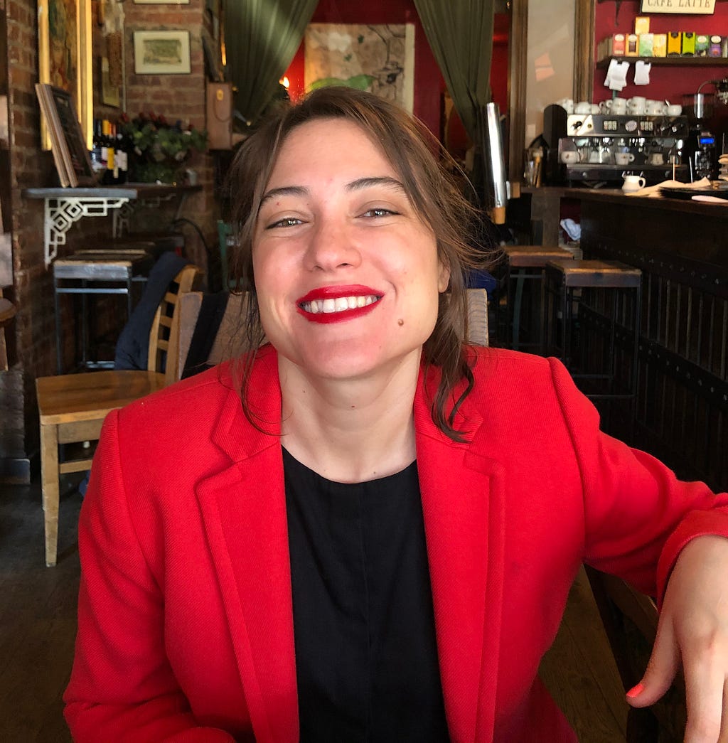 Cristina Pérez Díaz wears a red blazer and a black top with red lipstick. She has shoulder length brown hair, and is smiling.
