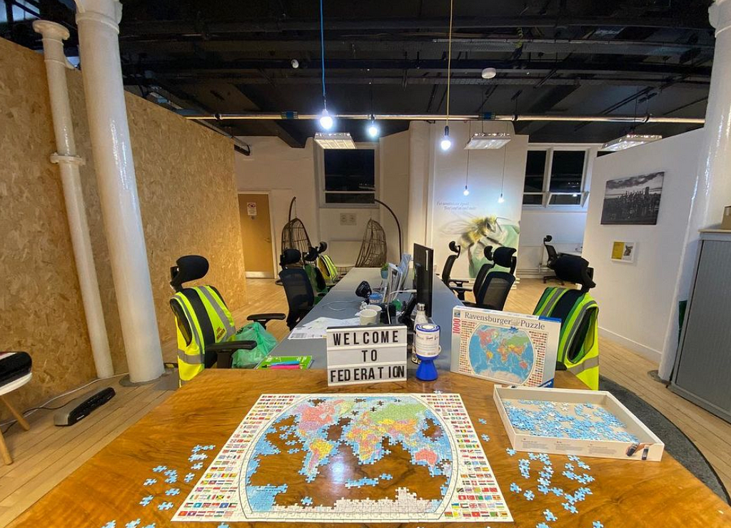 In the foreground is a partially complete jigsaw featuring a map of the world. It is laid out on a wooden desk in front of a lightbox with black lettering that reads: “Welcome to Federation”. In the background are co-working desks.