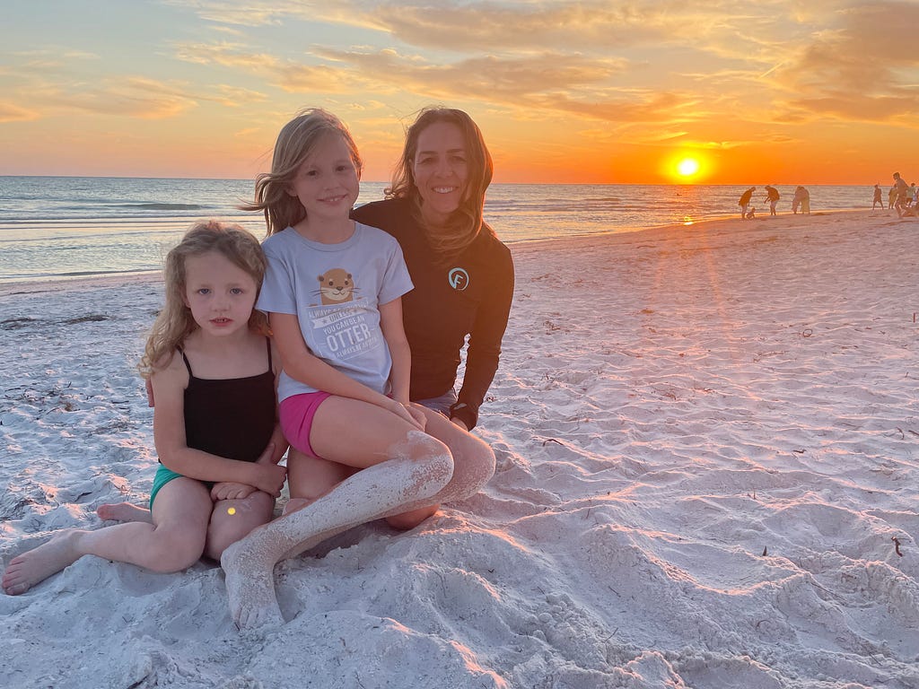 A mom and her two daughters sit in the sand as the sun sets behind them