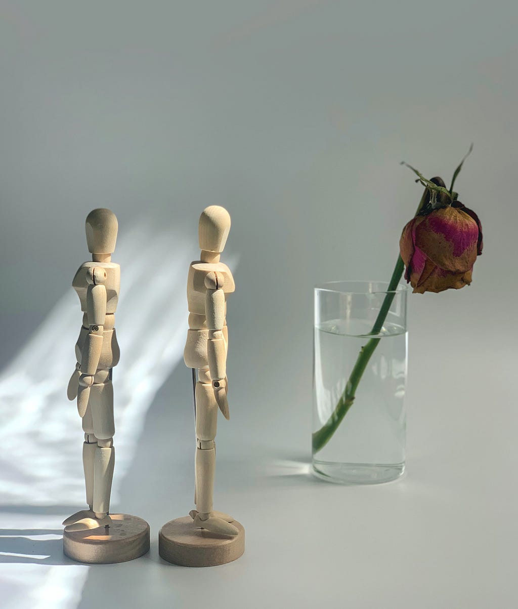 Two wooden form dolls face away from one another next to a wilting rose in a glass of water