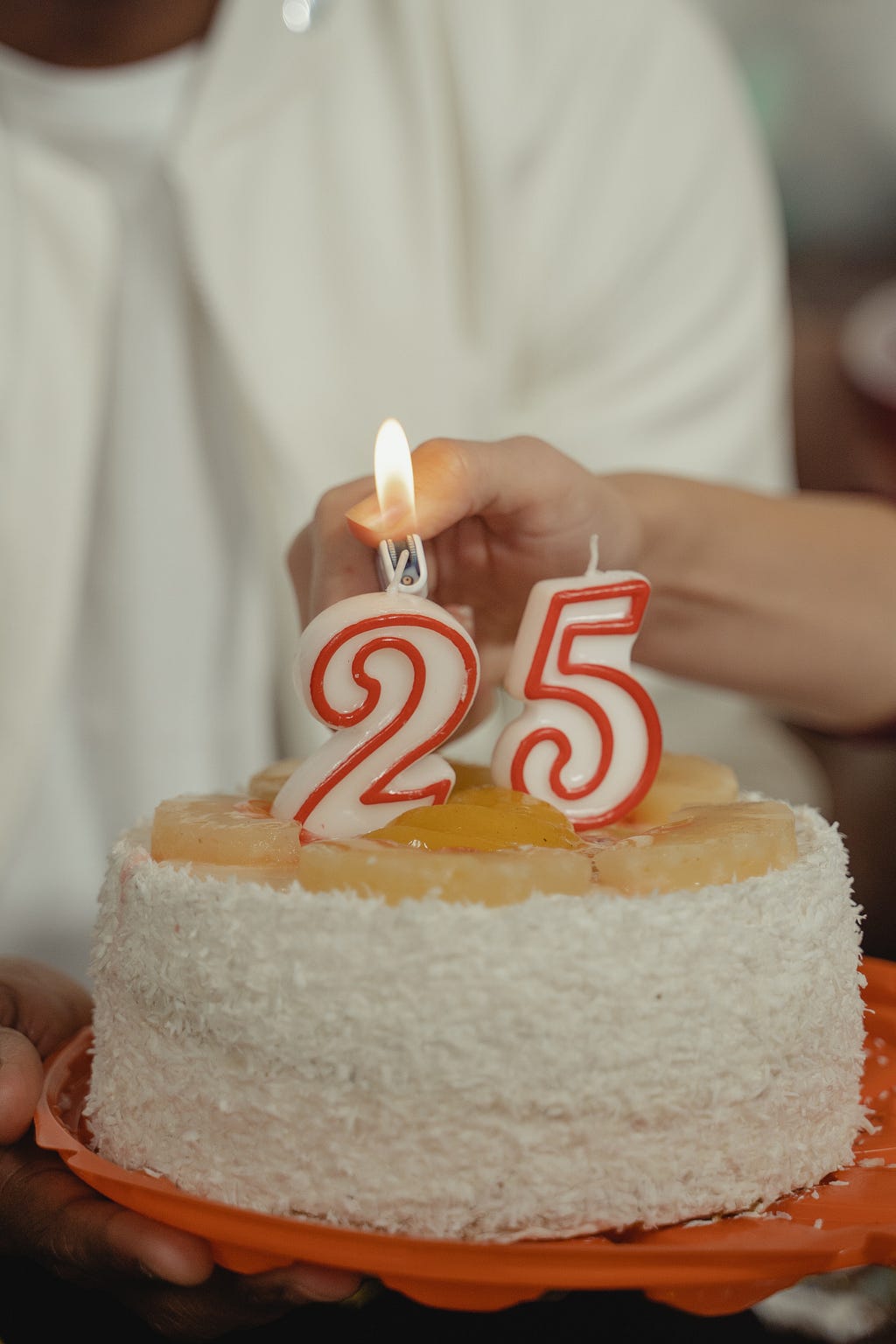 White Numbered birthday candles lined with red for the number twenty five sits atop a white coconut cake with a caramel top. The number two is being lit with a lighter.