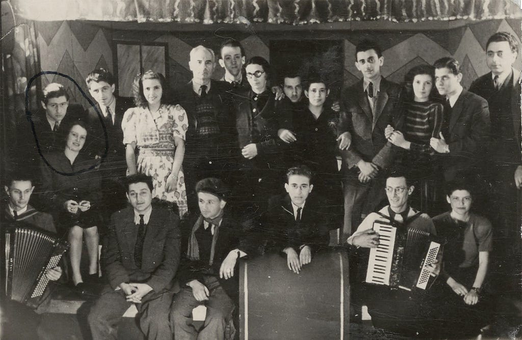 Nineteen adults in two rows, those in the back are standing and those in front are seated. Most of the men are wearing neckties. The women are wearing dresses or skirts. Some are smiling, linking arms, and leaning toward each other. At left, someone has drawn a circle around a couple. Two men in the front row hold accordions, one at far left and the other second from right.