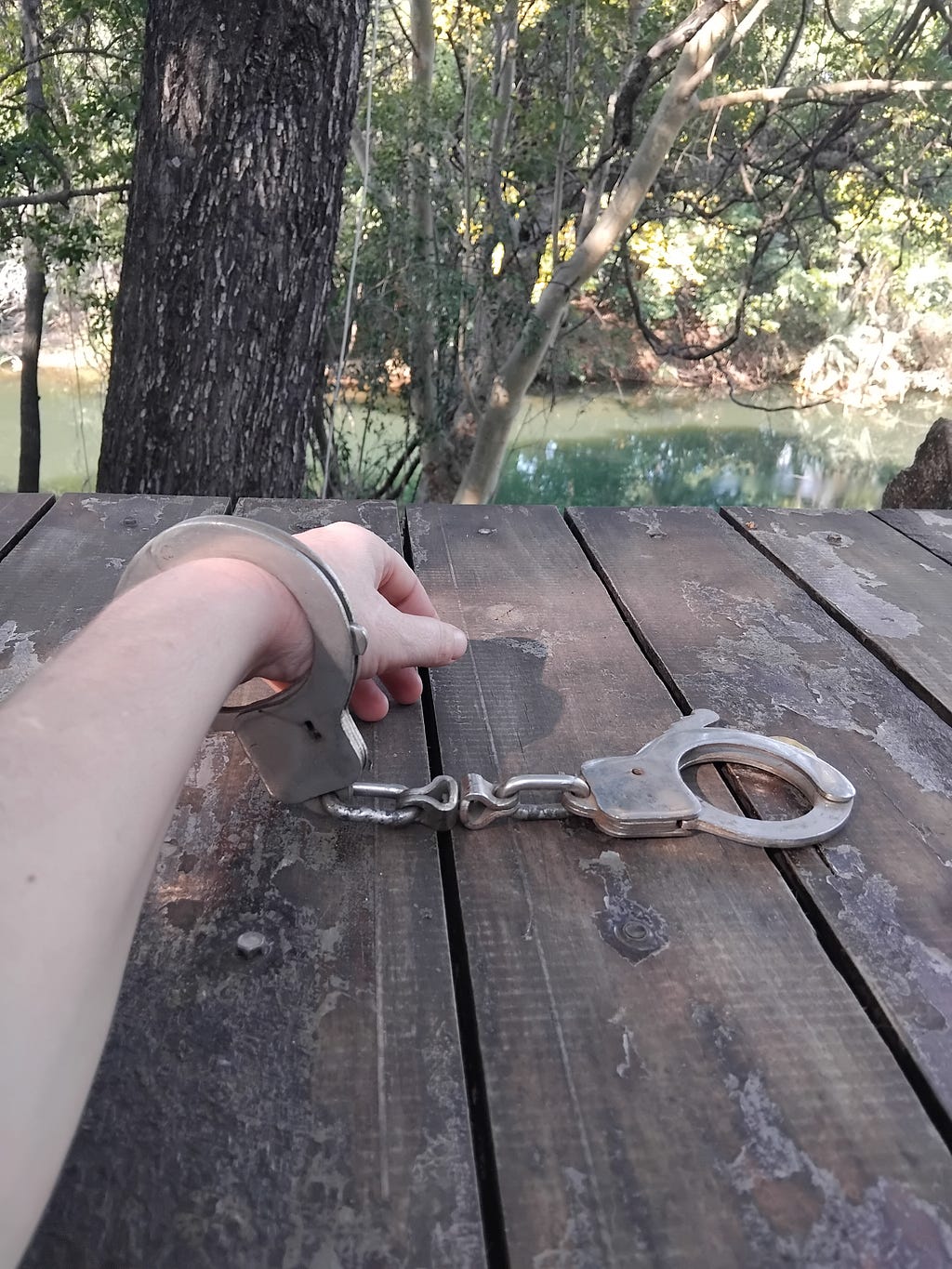 A Caucasian arm with one handcuff around the wrist, resting on a wood table, and a river in the background