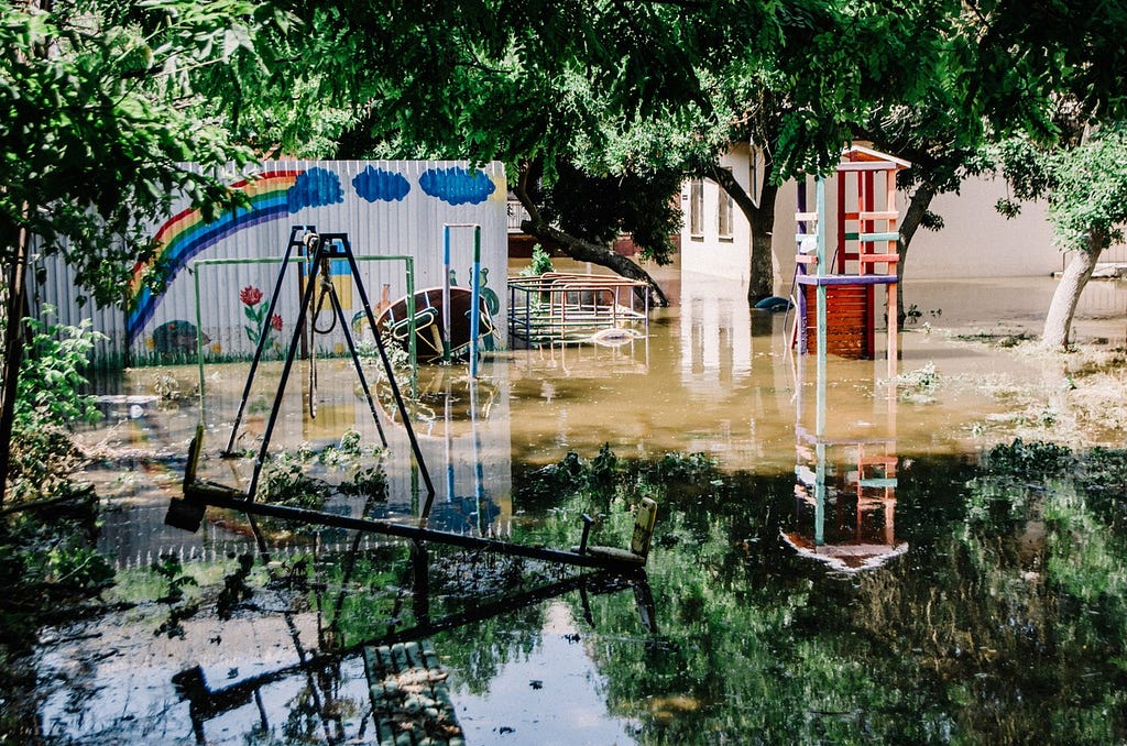 A flooded playground’s swing set and other equipment is reflected in the water that has partially submerged the area that is surrounded by still-green leafy trees.