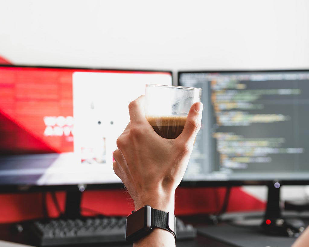 A white person’s hand holding a glass cup of coffee in front of two computer monitors