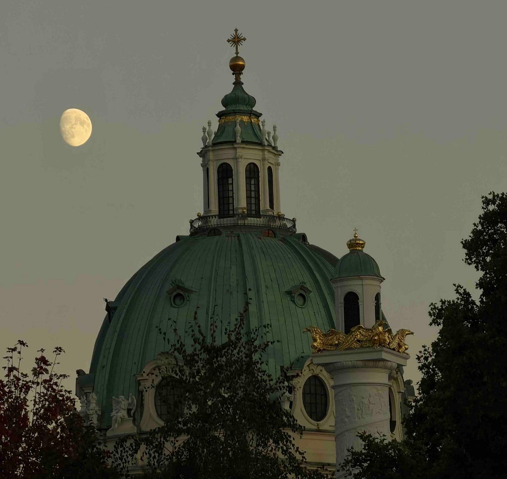 A grand domed building with classical architecture, featuring a green copper dome topped by a cupola and cross, stands partially obscured by trees. Ornate golden embellishments and statues accent the structure, especially on a turret to the right. A nearly full moon glows in the muted gray sky, suggesting dusk or dawn, adding a serene contrast to the building’s opulence.