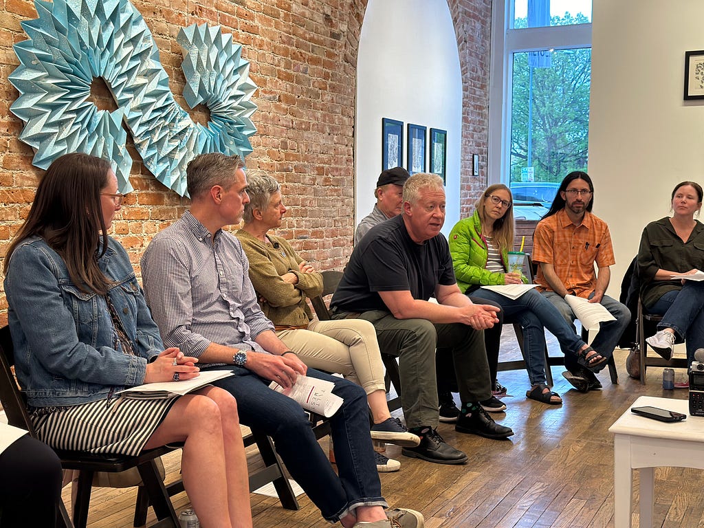 Eight people sit in a semi-circle as part of a larger group conversation. At their center is a man speaking who wears a blue t-shirt and dark blue pants. He has short grey hair. Around him are men and women listening. On the wall behind them are artworks, including one sculptural piece that appears to be a horizontal infinity symbol.