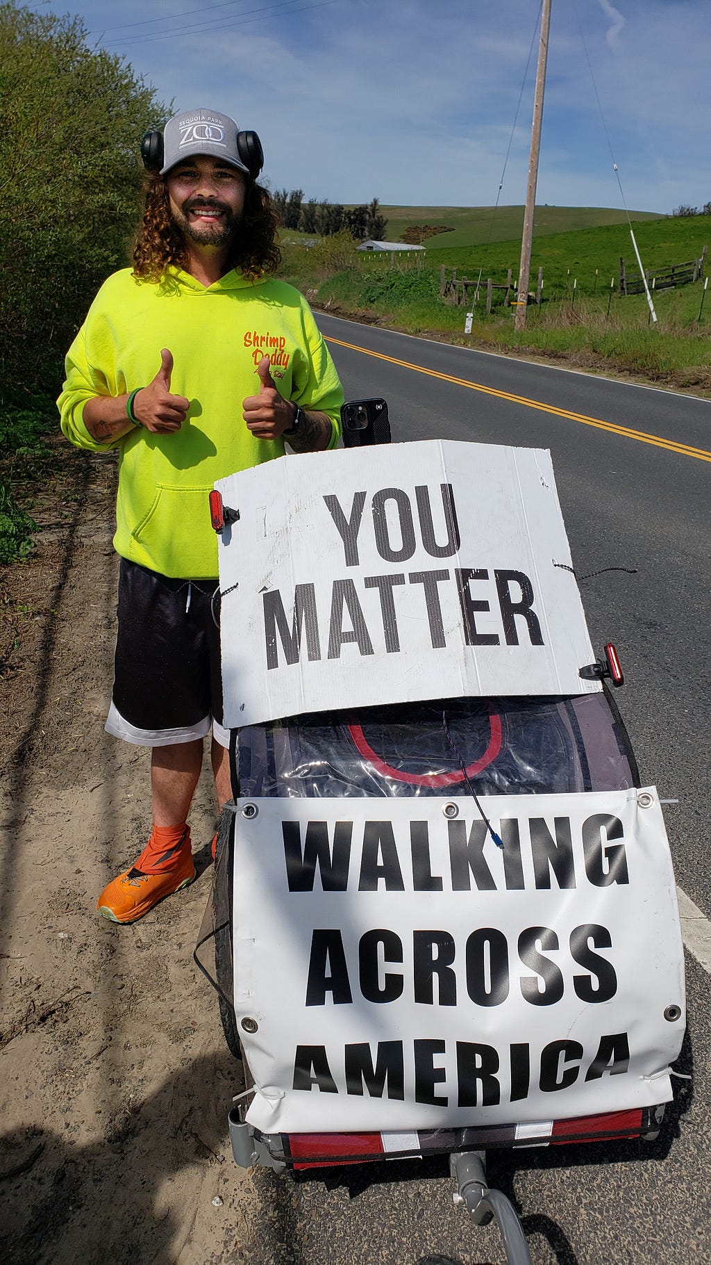 A young white man with curly, shoulder-length hair wearing a fluorescent green shirt, pushing a cart with his belongings and 2 signs — one says “You matter” and the other says “Walking across America.” He is standing on the dirt shoulder of a 2-lane country road.