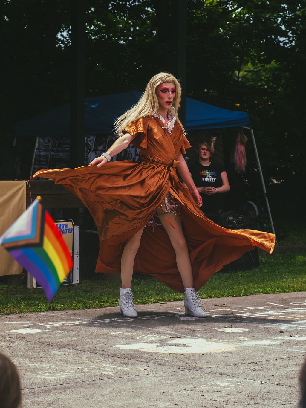 A drag queen spinning in an orange dress with white high heeled shoes & a white wig. A Pride flag can be seen in the foreground