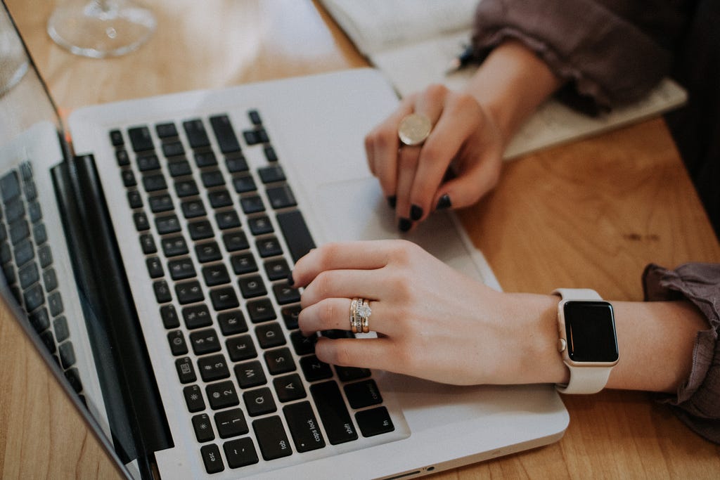 A person’s hands, wearing rings and a watch, are typing on a laptop.