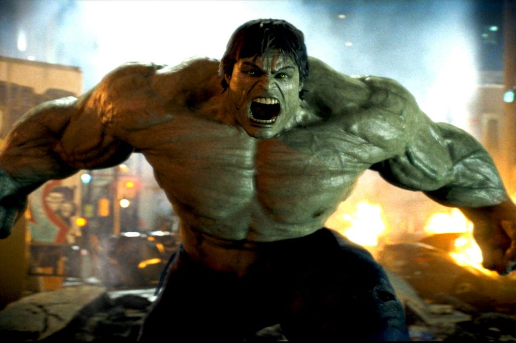 The Hulk, a large green monster with black hair, is screaming loudly with his muscles bulging. Behind him, a blurry city in disrepair.