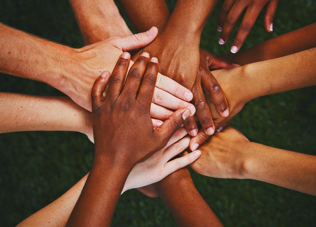 Many human hands coming together in a center touching. Green grass background under them.