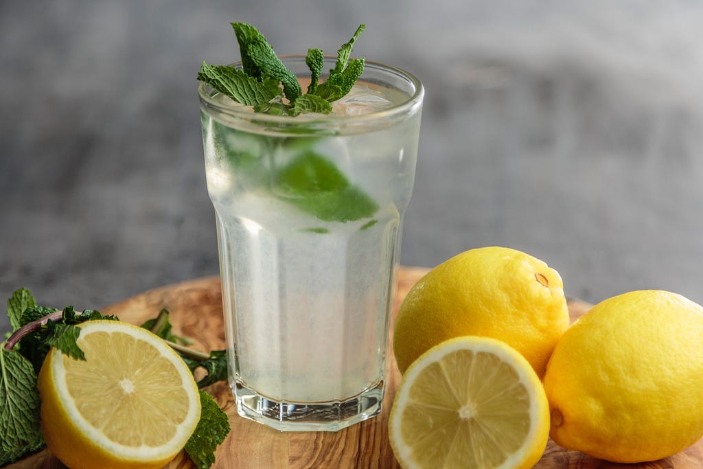 A glass with lemon juice and water, ice cubes and mint. Lemons are spread around it.