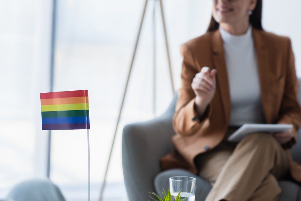 A psychologist holds a pen and paper in the background. The foreground shows a small Pride flag and a glass of water.l