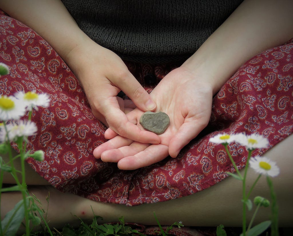 A small grey rock held on the palm of a white woman’s hands. Her hands are resting on her red paisley skirt and her legs can barely be seen tucked beneath her. There are little flowers in the foreground.