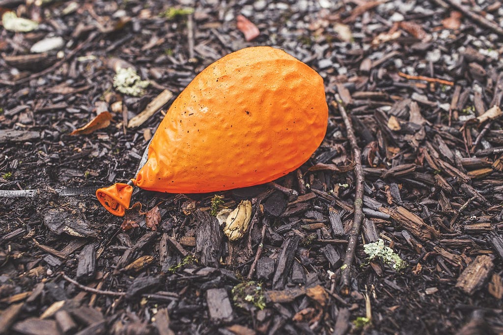A deflated orange balloon laying alone on damp woodchip ground