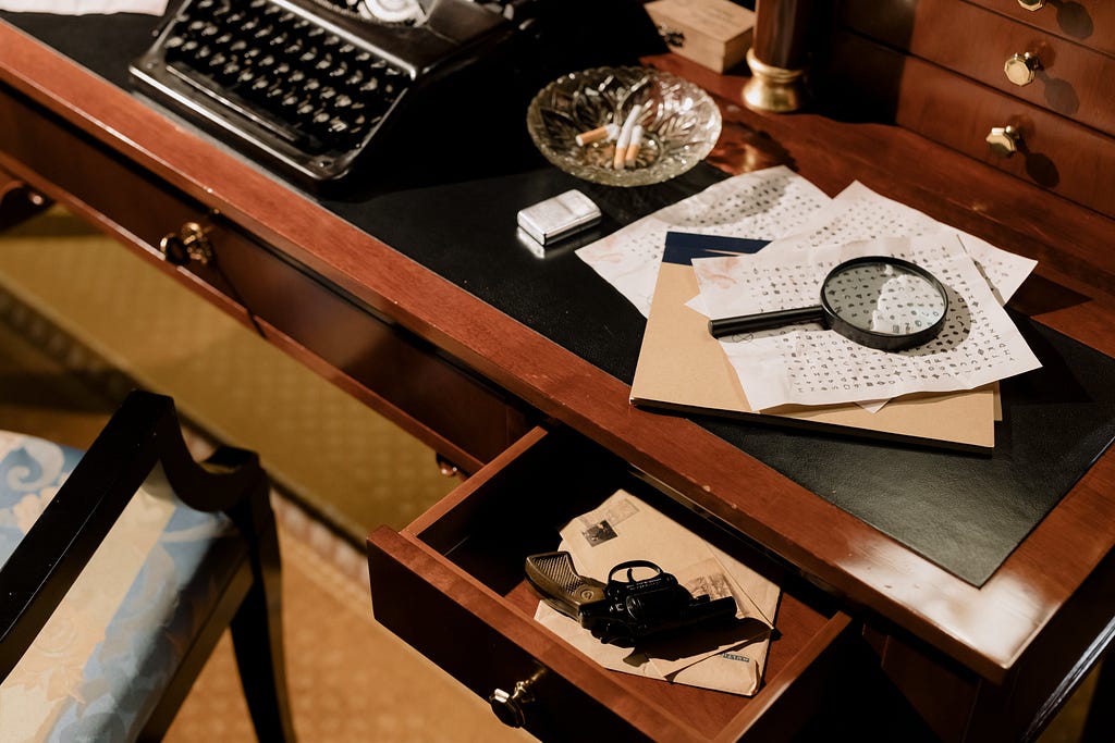 A 1940s-era desk with spy and investigator tools — typewriter, magnifying glass laid on a cipher, and a gun in an open desk drawer.