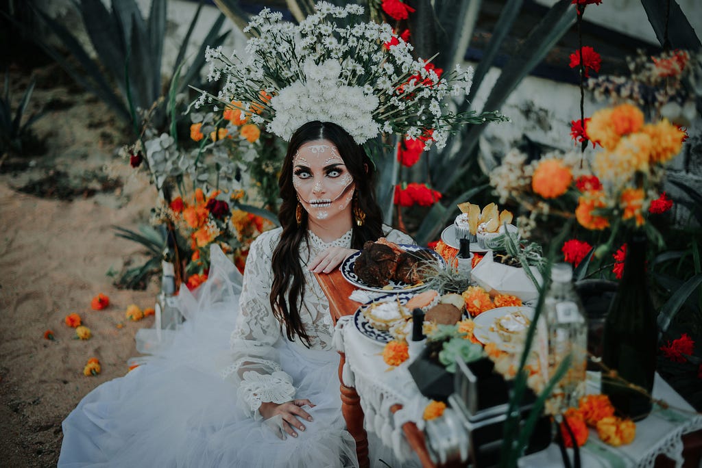 A Woman in Catrina Makeup Sitting beside an Altar with Food and Flowers