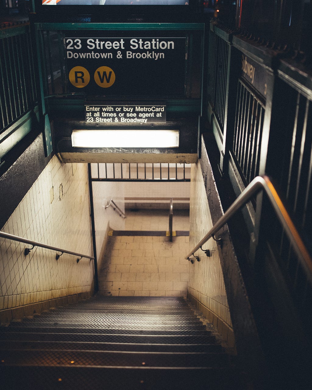 The entrance of a New York subway station with a sign indicating which lines pass through this station and additional subtext providing instructions about Metro card usage.