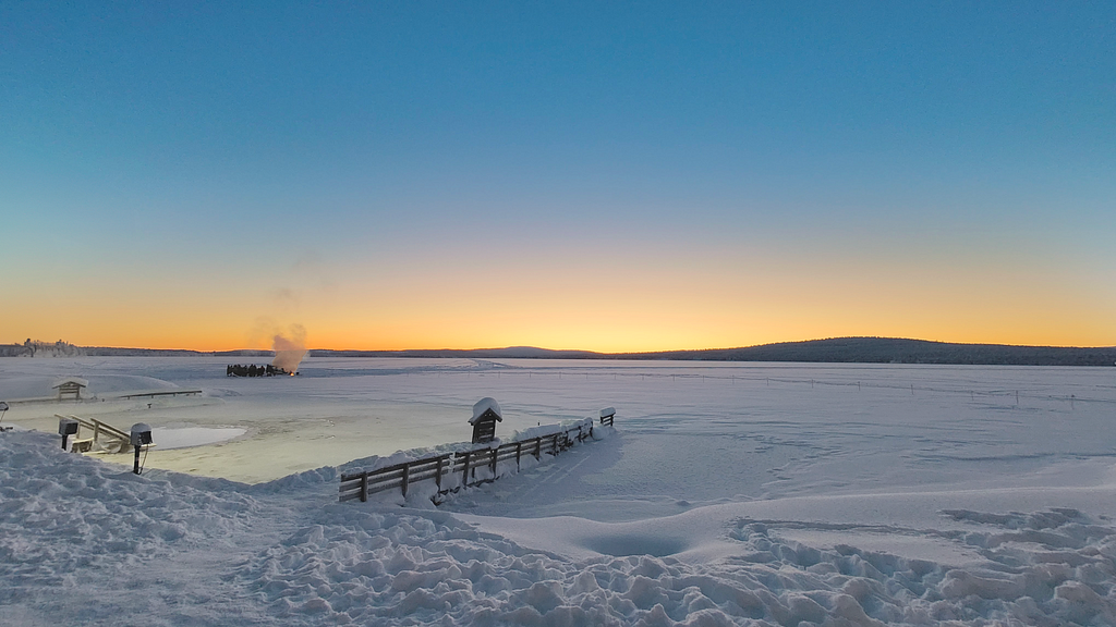 A rich sunset sets over a frozen lake that is covered in snow. In the foreground a hole is cut out of the lake, with water showing through. Just behind, a group of people gather around a small smoking fire.