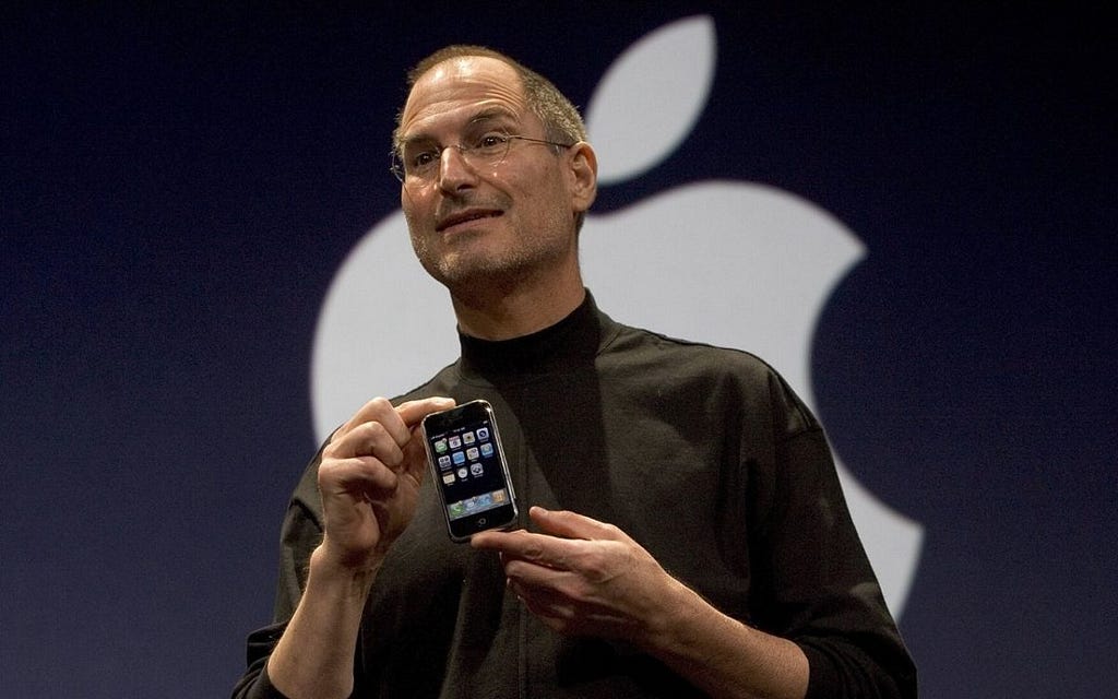 Steve Jobs former CEO of Apple holds the first iPhone in his hands in front of a large Apple logo behind him. Image taken during the unveiling in 2007.